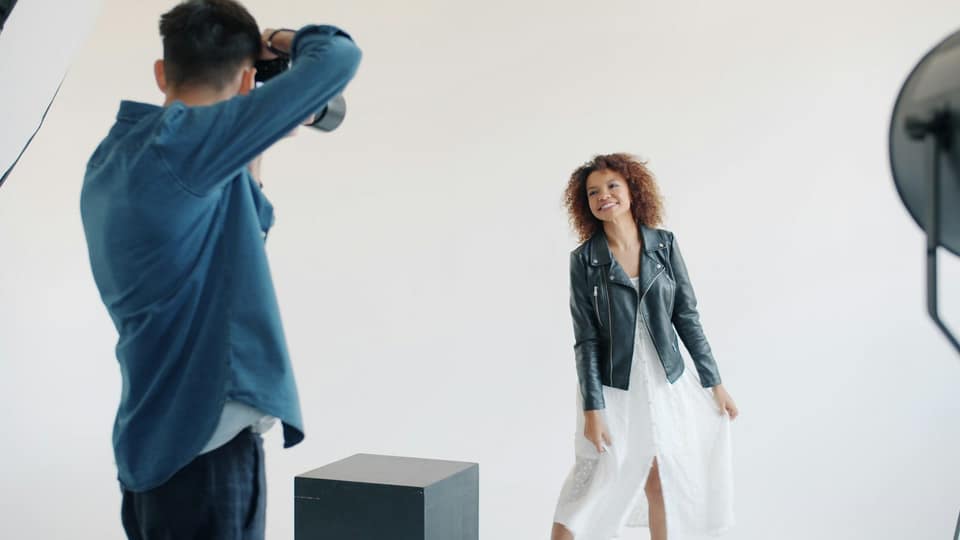 Een lachende vrouw met krullend haar in een leren jas en witte jurk poseert in een witte fotostudio, terwijl een fotograaf in een spijkerblouse haar fotografeert