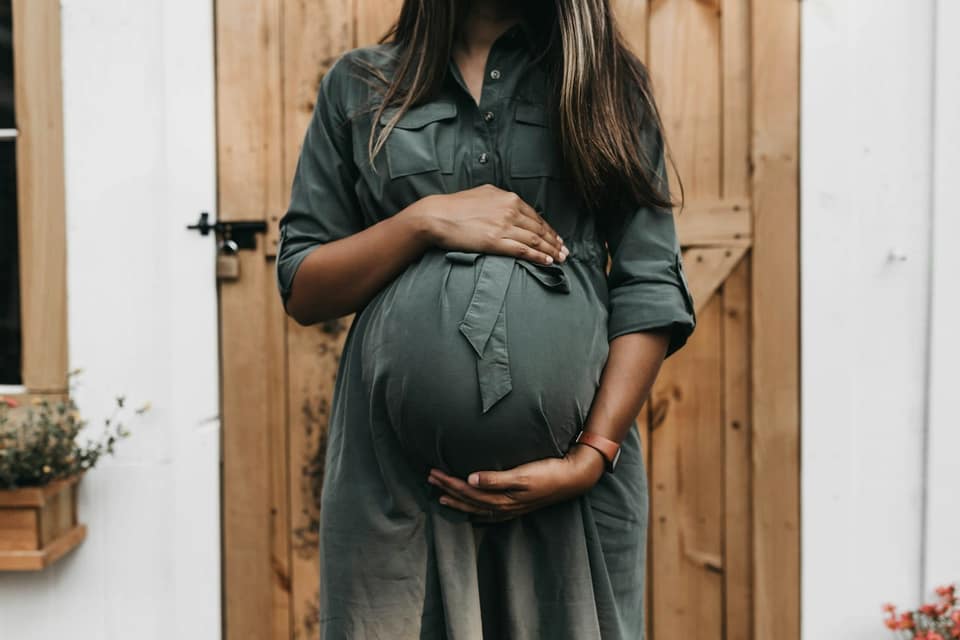 Zwangere vrouw in een groene overhemdjurk houdt haar buik vast, voor een houten deur, tijdens een zwangerschapsshoot