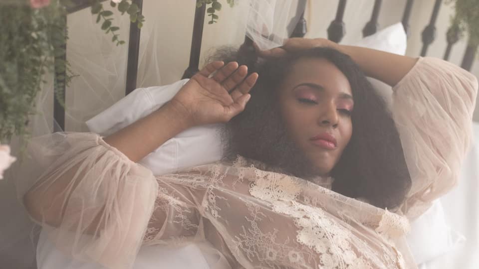 Een vrouw met lang donker krullend haar ligt met gesloten ogen op een wit kussen tegen een zwart ijzeren bed, in een roze kanten peignoir met tule mouwen