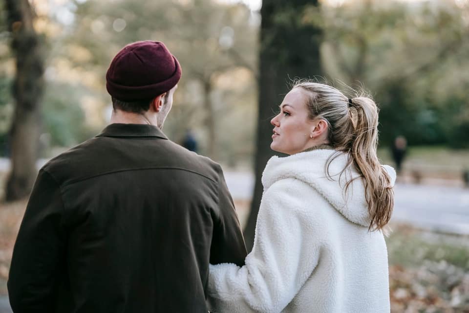 Een man met een rode muts en een vrouw in een witte teddyjas lopen arm in arm door een park in de herfst